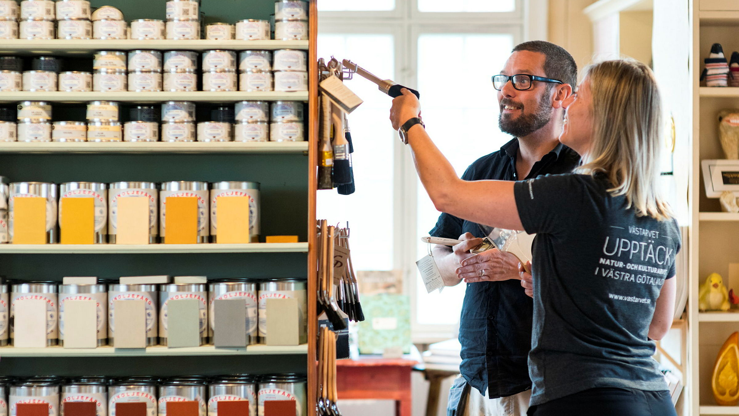 Två personer står vid en hylla med penslar och färgburkar i en äldre sekelskiftes-butiksmiljö.