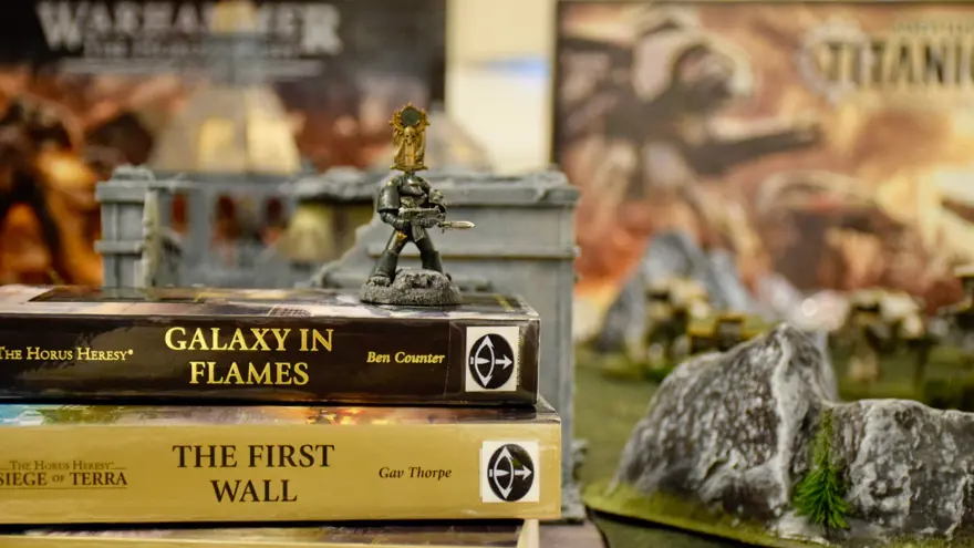 En spelfigur står på en hög med böcker. Böckerna har engelska titlar. Bredvid finns en bergformation som tillhör spelet.