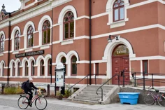 Tidaholms bibliotek, en röd byggnad med vita fönster som är formade som bågar längst upp. En kvinna cyklar förbi, på huset står skylten TIDAHOLMS BIBLIOTEK.