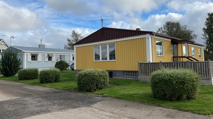 Ett par låga enplanshus, det främre gult och det bakre ljust gråblått. Rundklippta buskar framför husen. Bakom syns den blå himlen med vita molntussar.