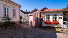 En gata med kullersten och låga äldre trähus. En kvinna med rullator står utanför ett av husen, ett annat hus har stora röda pelargoner i blomlådor vid fönstren. Solen skiner.