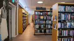 Bokhyllor i ljust trä fyllda med böcker står i biblioteksrummet. Väggen är en tegelvägg, en stor svartvit bild av en äldre kvinna i vitt förkläde och runda glasögon pryder väggen.