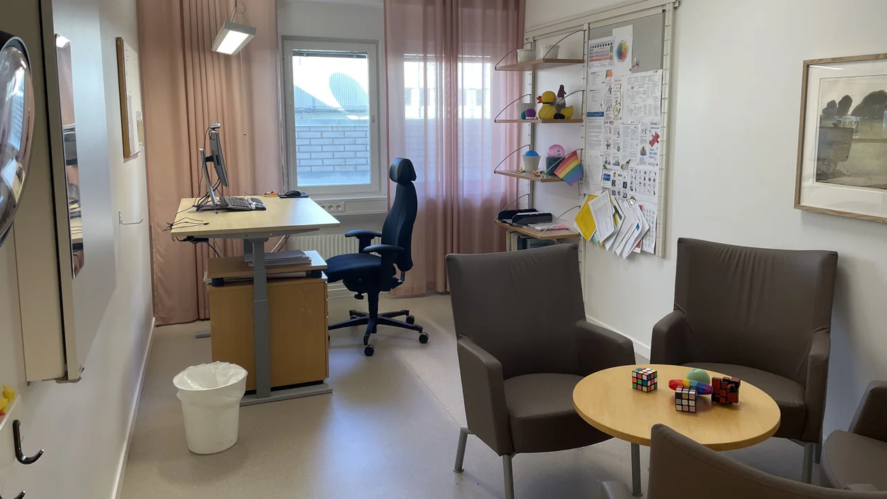 Behandlingsrum med grå fåtöljer vid ett runt litet bord. Längre bort i rummet finns ett skrivbord med en dator och en kontorsstol.