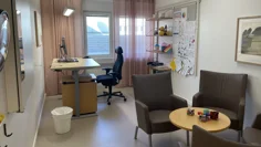 Behandlingsrum med grå fåtöljer vid ett runt litet bord. Längre bort i rummet finns ett skrivbord med en dator och en kontorsstol.