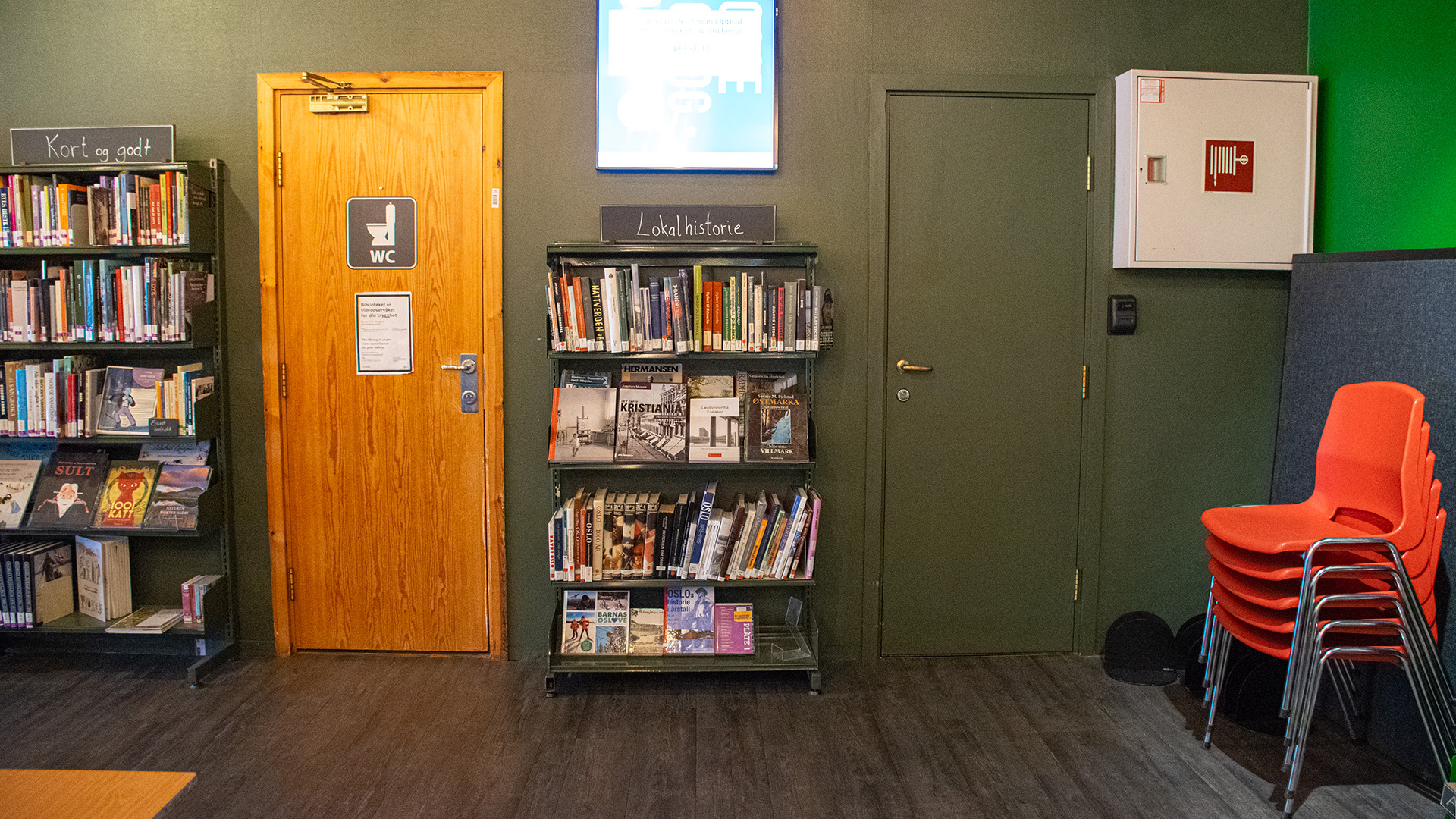 Två dörrar på samma vägg. Väggen är målad i en militärgrön färg, den ena dörren är målad i samma färg, den andra är omålad. Den omålade dörren har en WC-skylt på sig. Mitt emellan dörrarna finns en låg bokhylla med skylten "Lokal historie". .