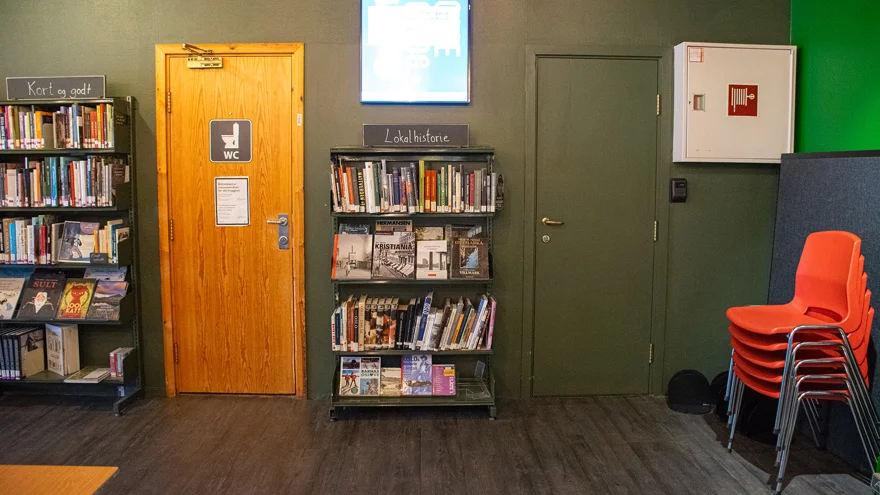 Två dörrar på samma vägg. Väggen är målad i en militärgrön färg, den ena dörren är målad i samma färg, den andra är omålad. Den omålade dörren har en WC-skylt på sig. Mitt emellan dörrarna finns en låg bokhylla med skylten "Lokal historie". .