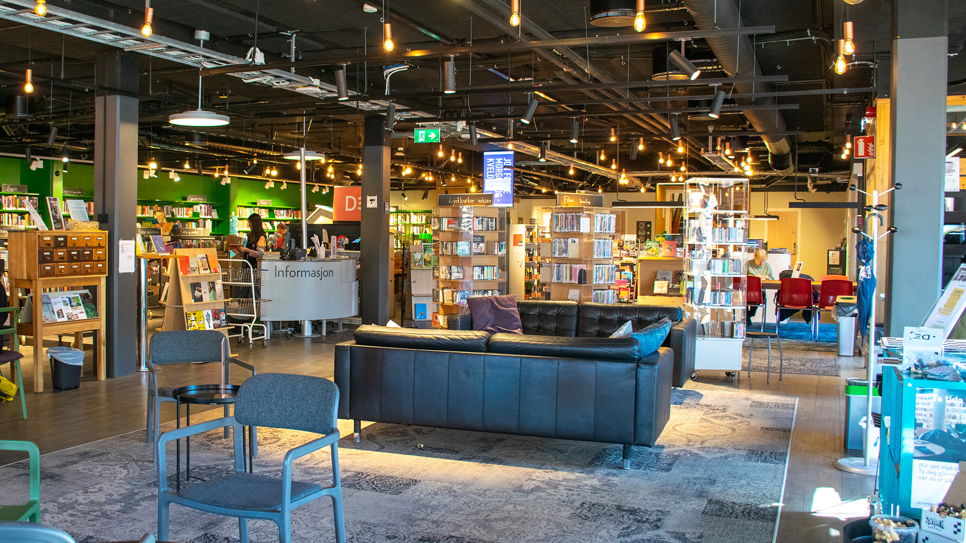 En soffgrupp i svart skinn står i biblioteksrummet. En stor matta ligger på golvet och i taket hänger små spotlights. Informationsdesken finns till vänster om soffgruppen och entrén till biblioteket finns till höger om den.  