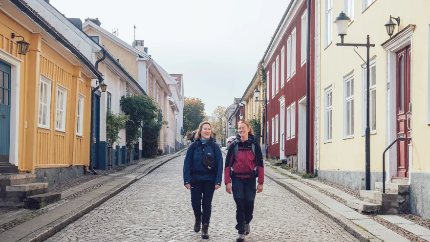Två kvinnor passerar färgglada trähus på en kullerstensgata.
