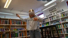 En flicka står med VR-headset inne bland bokhyllorna på barn- och ungdomsavdelningen på biblioteket i Kungshamn. Hon spelar och är i rörelse, i händerna håller hon spelkontroller. Hon har långt blont hår och är klädd i blå jeans och en ljusblå blus.