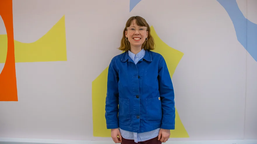 Rachel Barron är klädd i en ljusblå skjorta och en mörkblå jacka. Hon har ljusbrunt hår till axlarna och bär glasögon. Hon står mot en vit vägg med stora mönsterbitar i gult, orange och ljusblått.