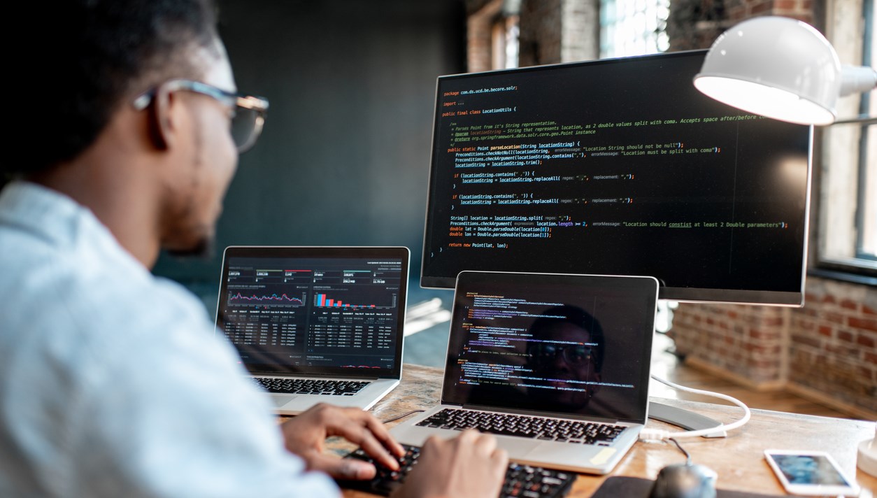 Young african male programmer writing program code sitting at the workplace with three monitors in the office. Image focused on the screen