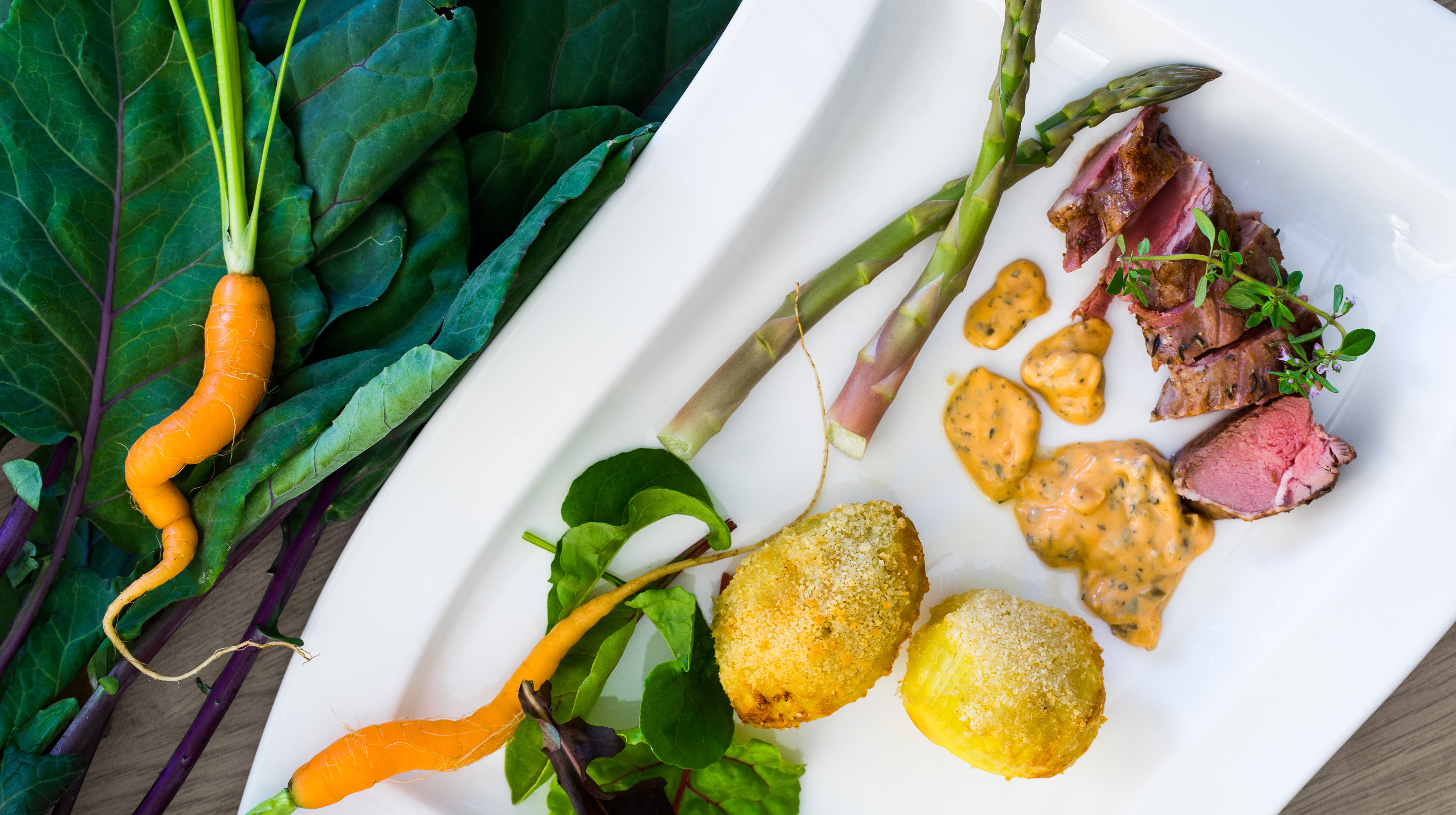 Vit tallrik med sparris, kött och potatis med gröna blad och morot bredvid