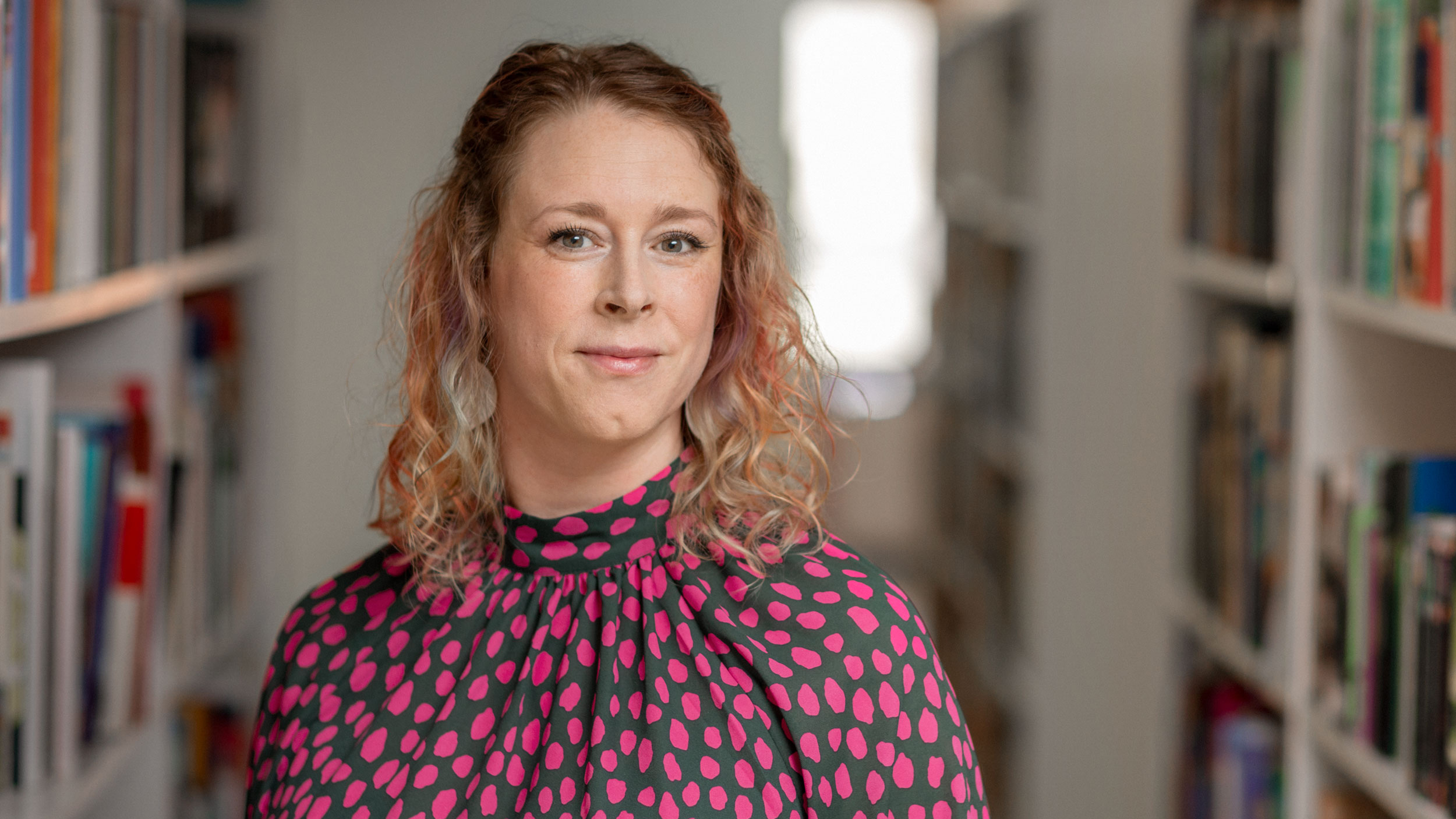 En kvinna med mörkblont hår står framför bokhyllor. Hon har på sig en prickig blus i blått och rött. 