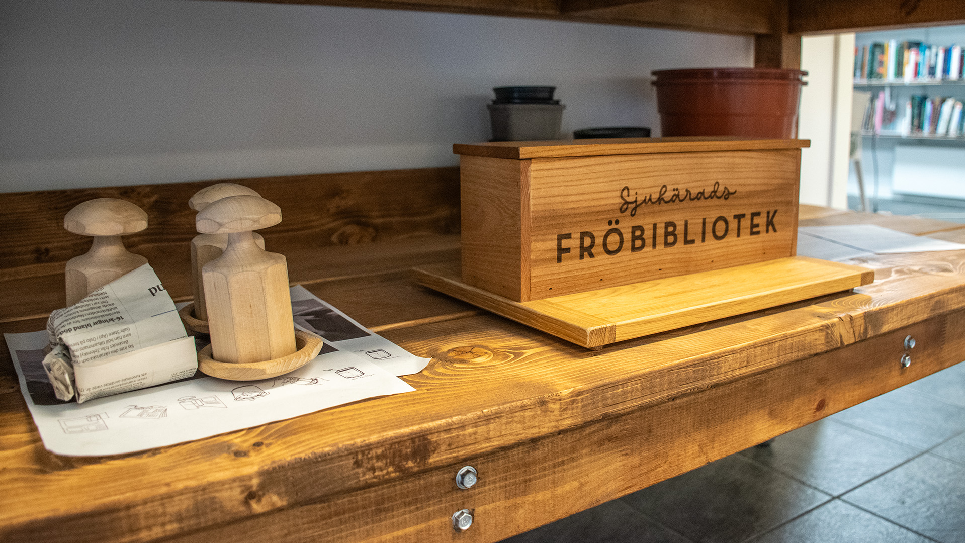 En hylla i trä, på den finns ett träverktyg som man kan göra odlingsurnor i tidningspapper med. En stor trälåda med texten Sjuhärads fröbibliotek och krukor i plast står också på hyllan.