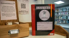 En gästbok med texten Fröbibliotek står på en bänk. På boken står instruktioner till hur man lånar fröer ut fröbiblioteket, Bokhyllor fyllda med böcker finns i bakgrunden. Bredvid boken står en trälåda med texten Annuell Bienn.