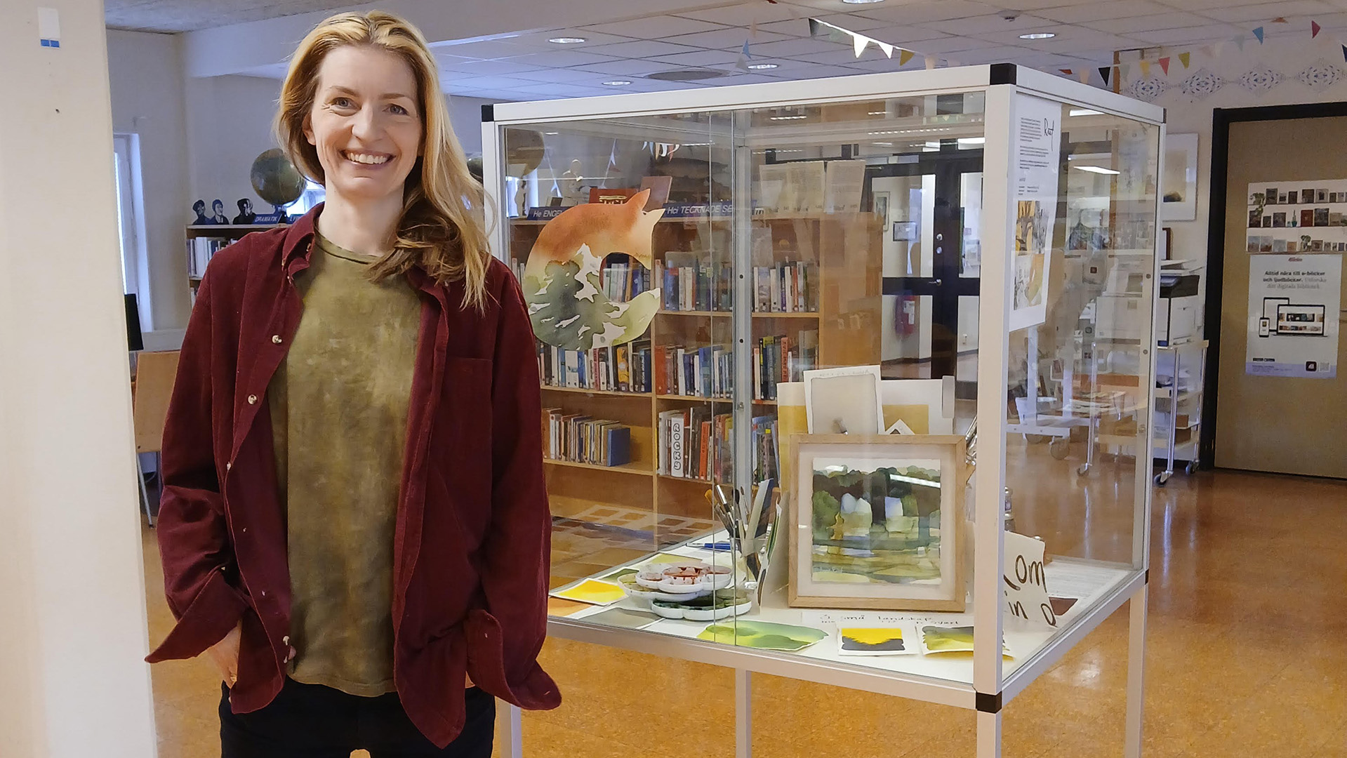 Michelle Fundin är i 40-årsåldern, hon har långt ljust hår och är klädd i en grön t-shirt med röd skjorta över. Hon ler stort mot fotografen, hon står inne på biblioteket i Ydre bredvid en glasmonter med målningar i.