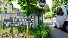 En golfbil susar förbi på en smal väg. Två träskyltar visar vägen, på den ena står det Bibliotek, på den andra Missionskyrkan.