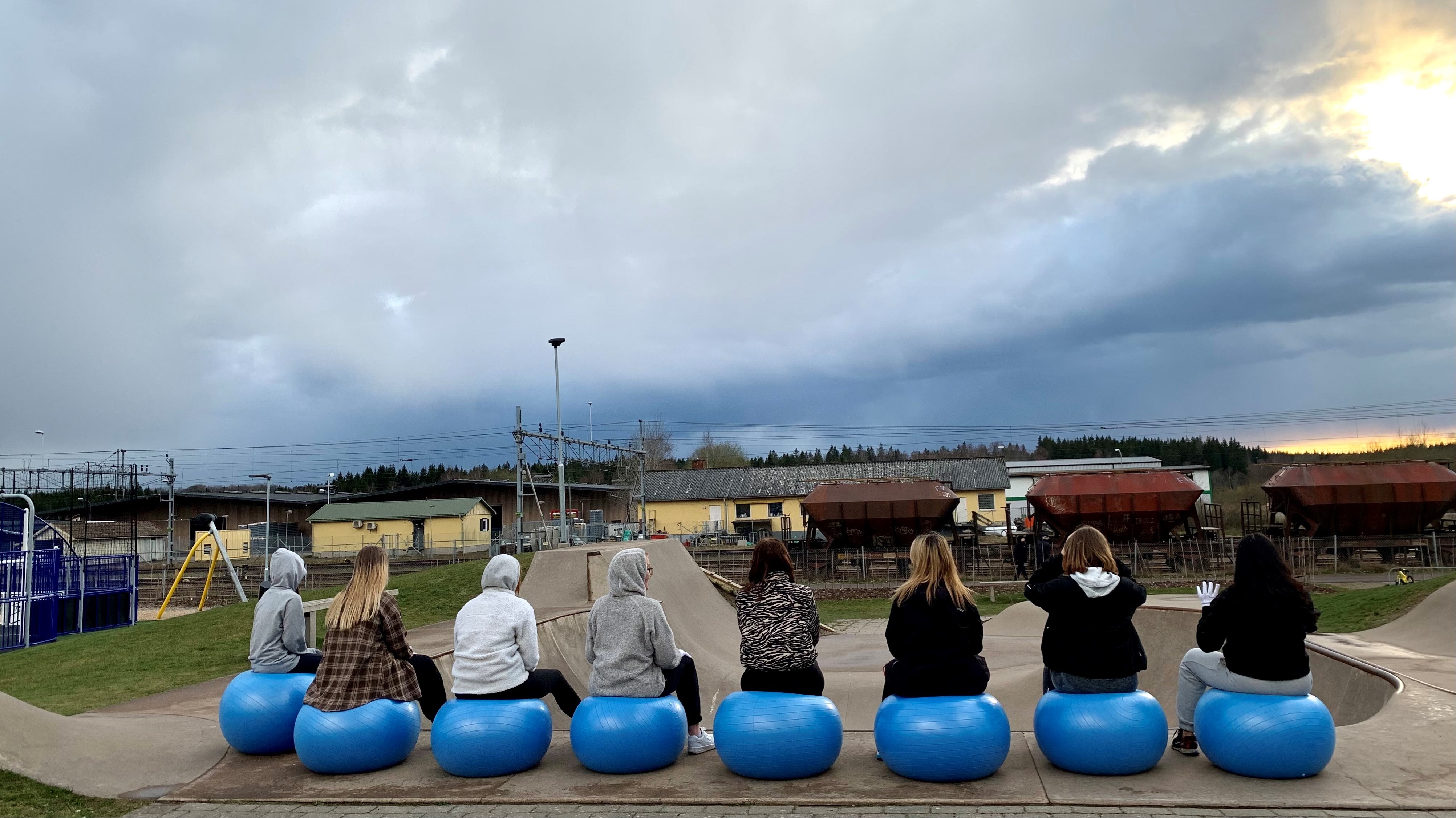 Åtta dansare som sitter på blåa pilatesbollar, utomhus, med ryggen åt kameran.