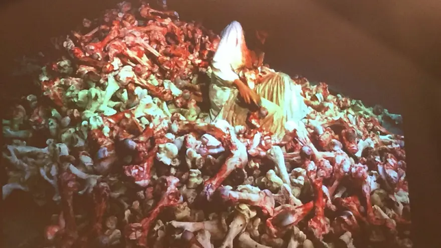 En människa i en vit särk sitter på en hög med blodiga ben.