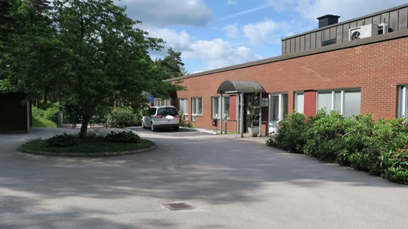 Låg röd tegelbyggnad med rund vändplats med träd i mitten framför.